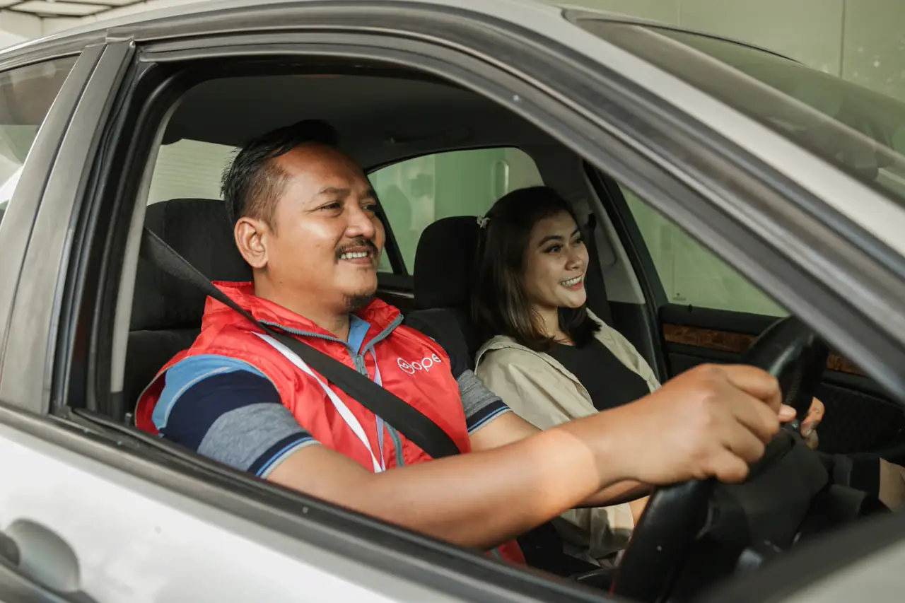 Homem dirigindo com uma passageira ao lado, os dois sorrindo.