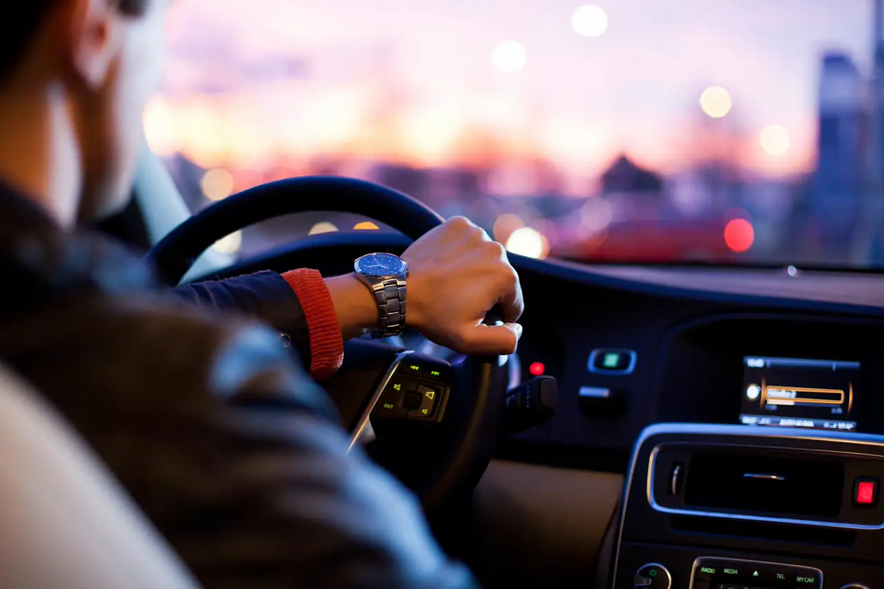 Homem dirigindo com uma das mãos no volante ao entardecer.