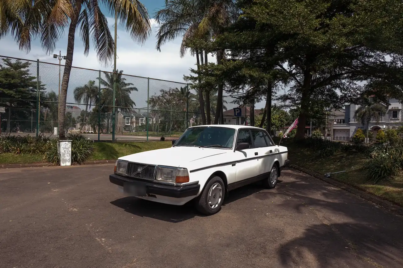 Sedã branco antigo parado num estacionamento cercado de árvores.