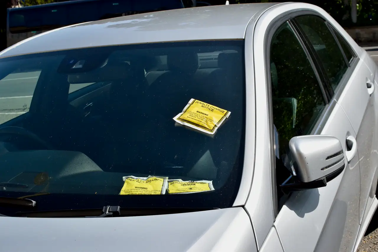 Carro prateado com avisos de multa amarelos no para-brisa.