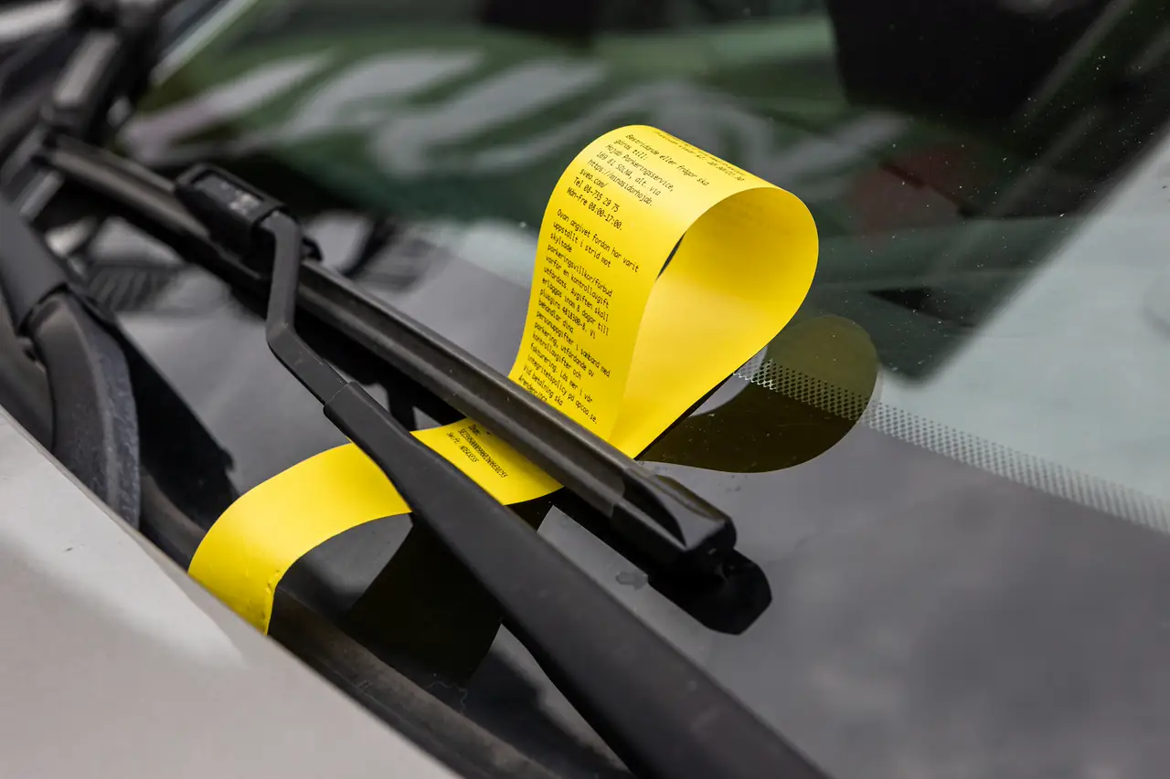 Notificação de multa amarela presa no limpador do para-brisa de um carro.