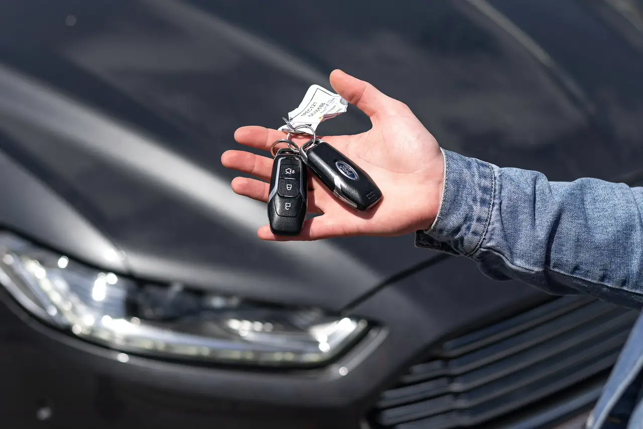 Mão oferecendo um chaveiro com duas chaves de carro ao lado de um carro escuro.