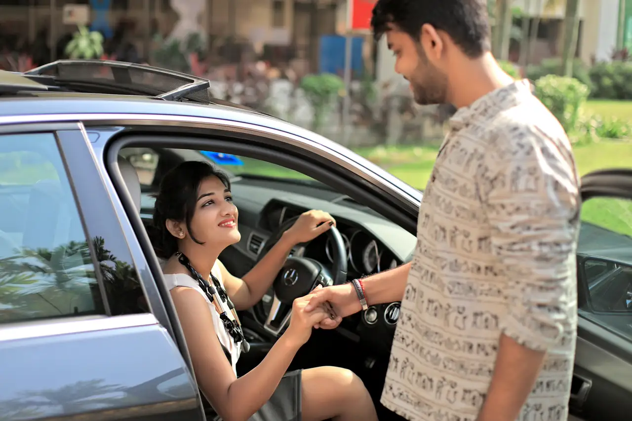 Homem cumprimentando a mulher sentada ao volante de um carro.