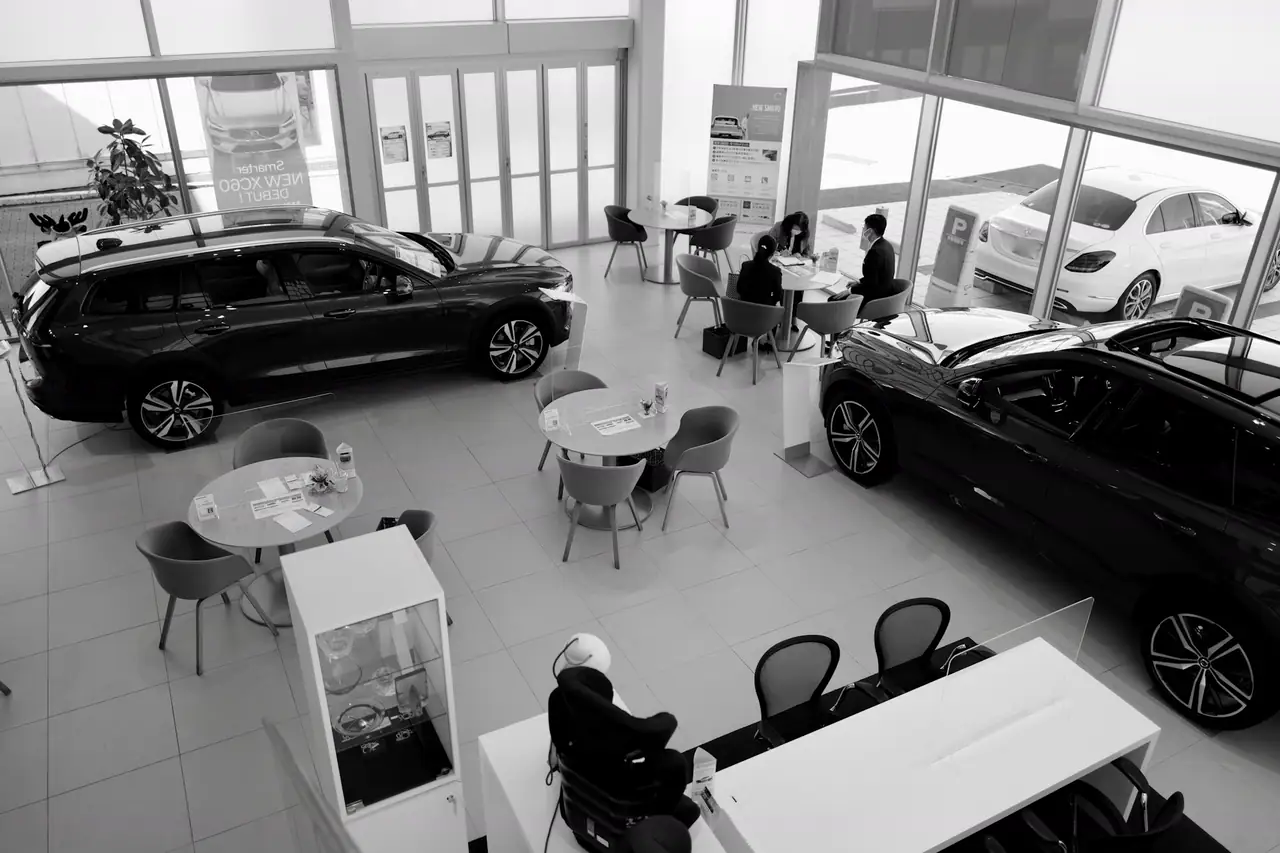 Salão de uma concessionária visto do alto, com carros expostos e clientes conversando nas mesas de atendimento.