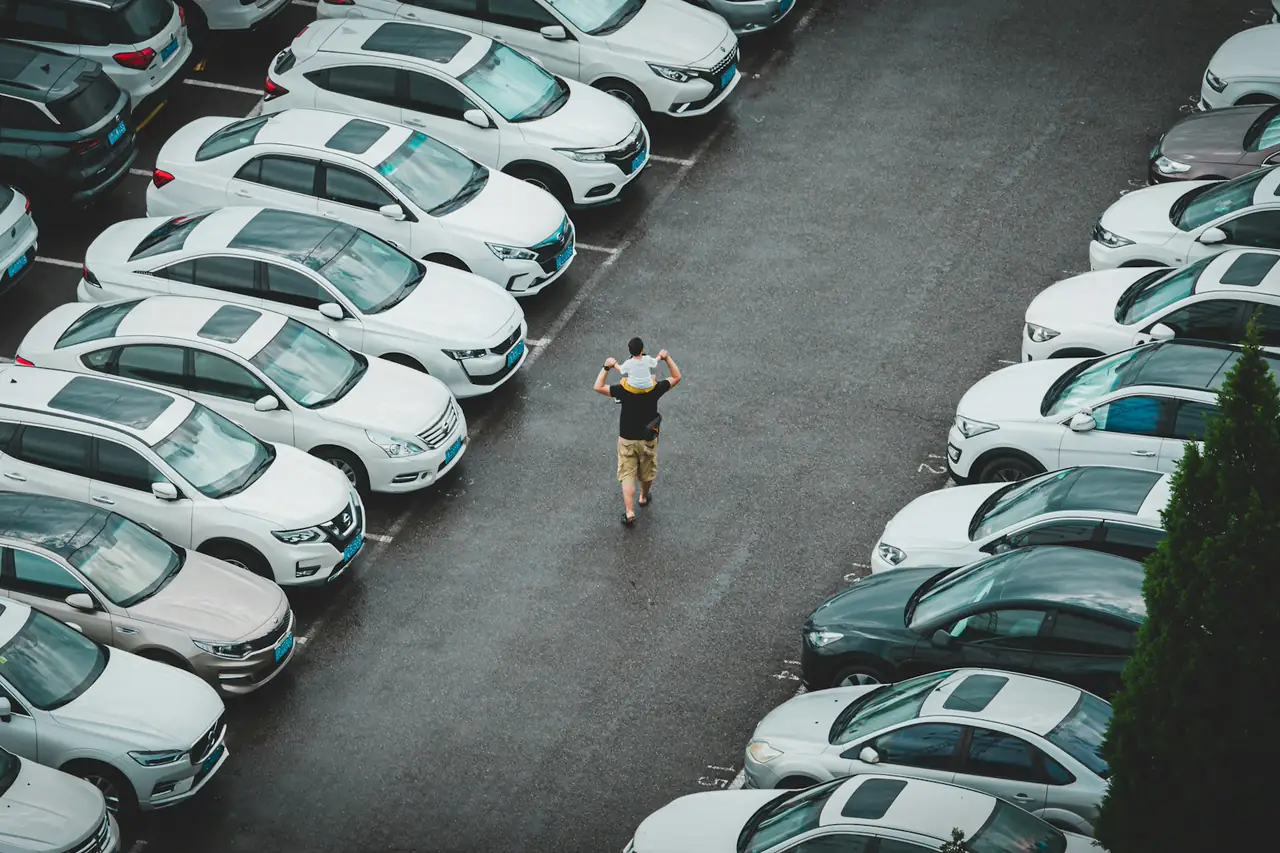 Pessoa caminhando entre duas fileiras de carros estacionados num pátio, vista do alto.
