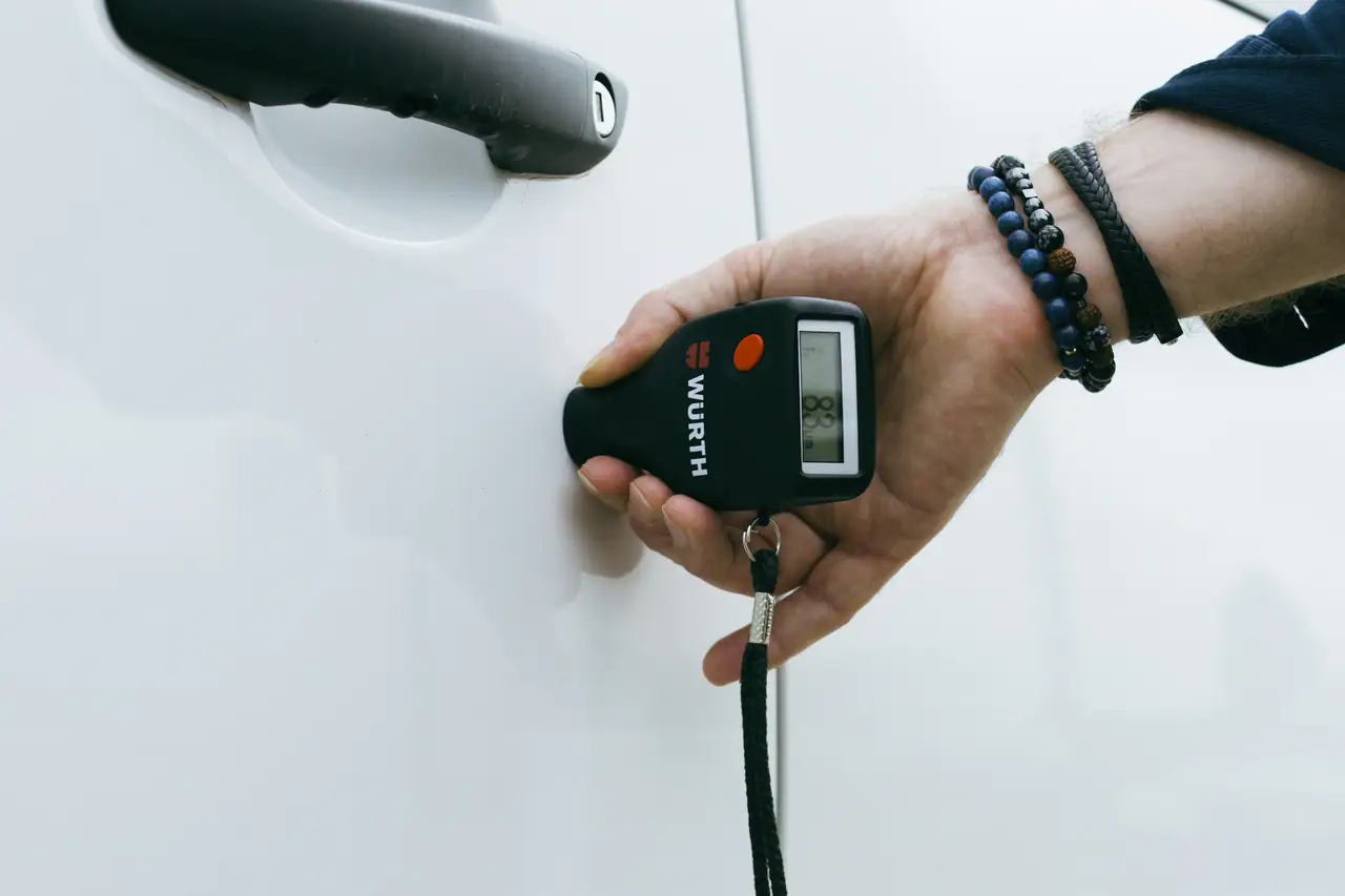 Mão segurando um medidor de espessura de pintura encostado na porta branca de um carro.