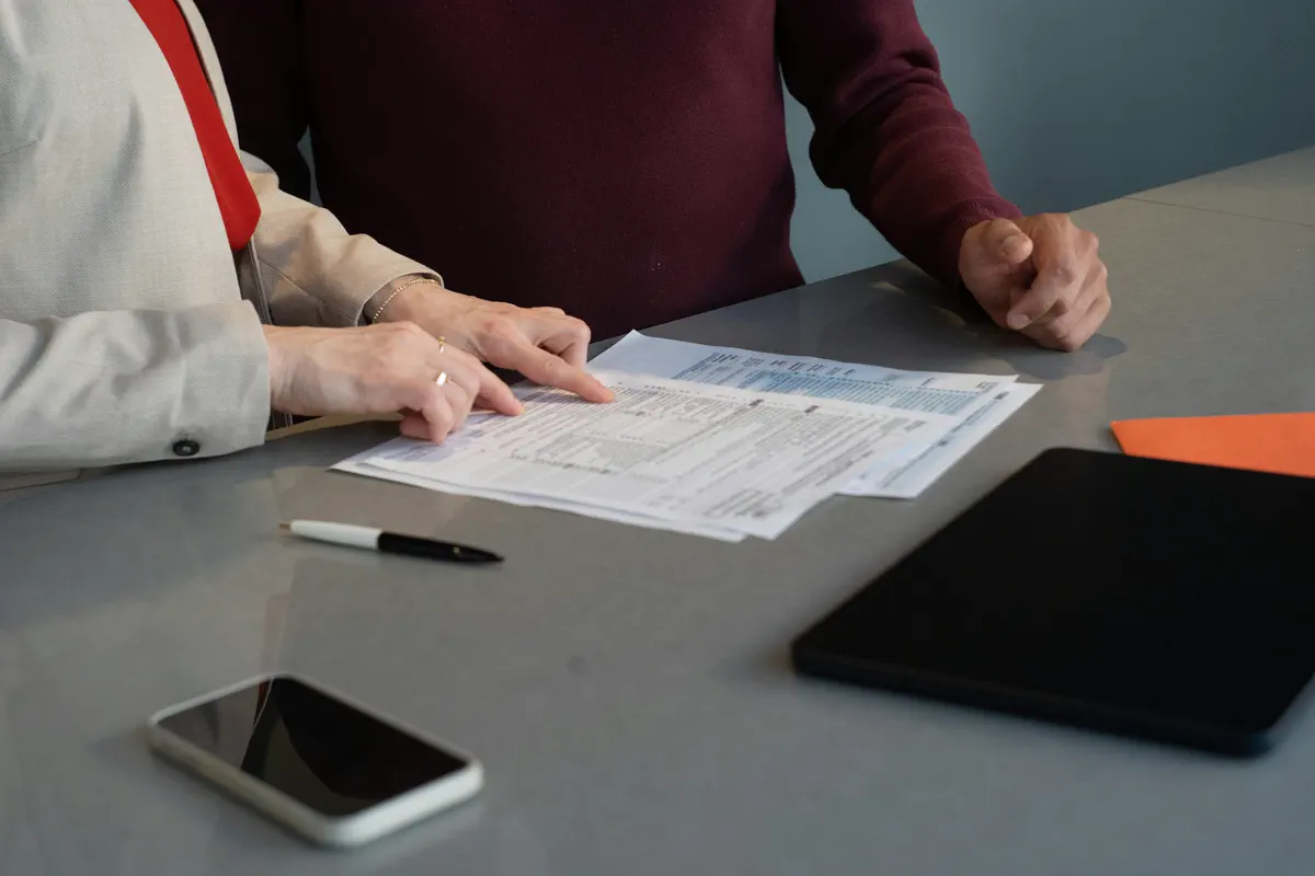 Duas pessoas conferindo formulários impressos sobre uma mesa, com celular e tablet ao lado.