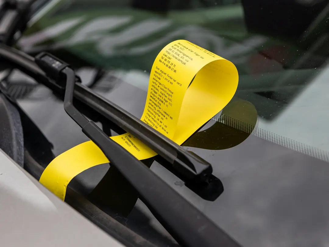 Notificação de multa amarela presa no limpador do para-brisa de um carro.