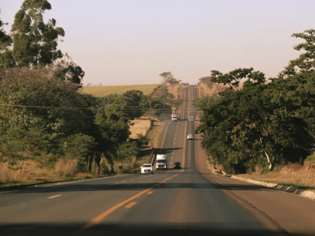 Rodovia de pista simples no interior do Brasil, com árvores dos lados e caminhões ao fundo.