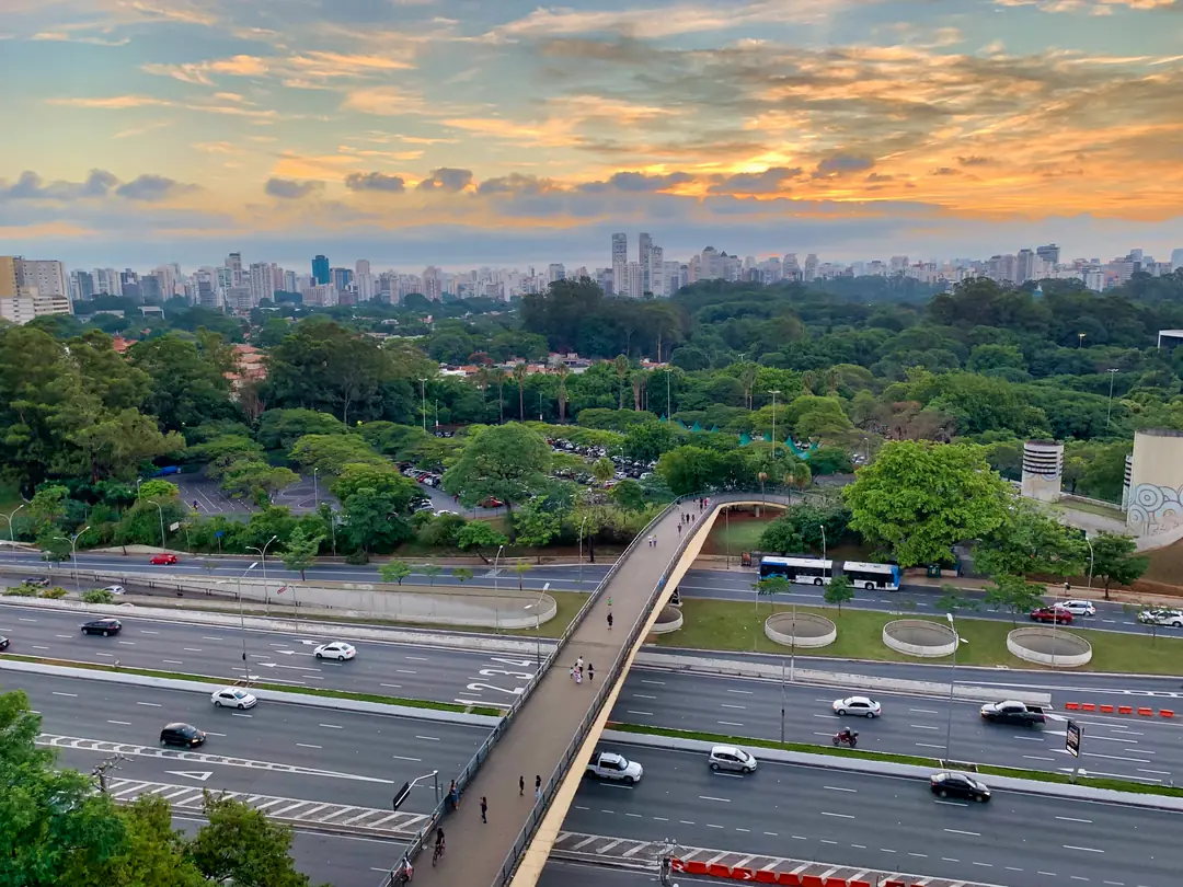 Vista aérea de uma avenida expressa de São Paulo com trânsito, parque e prédios ao fundo.
