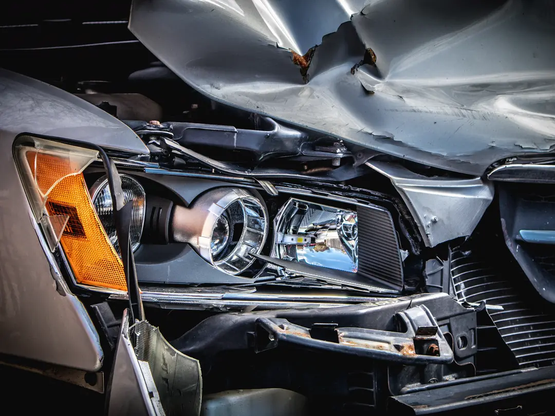 Frente de um carro prateado amassada após colisão, com o capô dobrado e o farol quebrado.